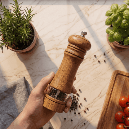 Hand holding a wooden pepper grinder with peppercorns on a marble surface, surrounded by potted plants and a cutting board with tomatoes.