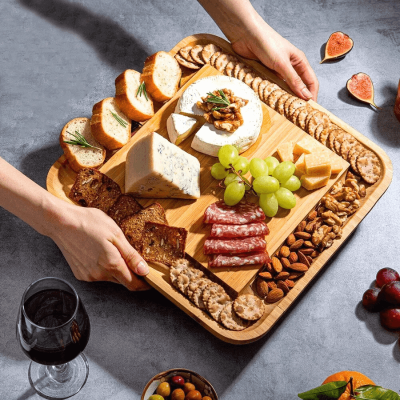 Wooden charcuterie board with assorted cheeses, meats, fruits, and nuts on a gray surface.