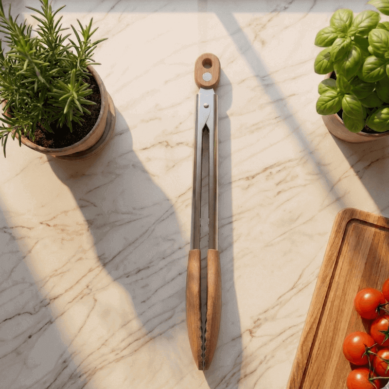 Woodastic grill tongs with wooden handles on a marble surface with potted plants and tomatoes.