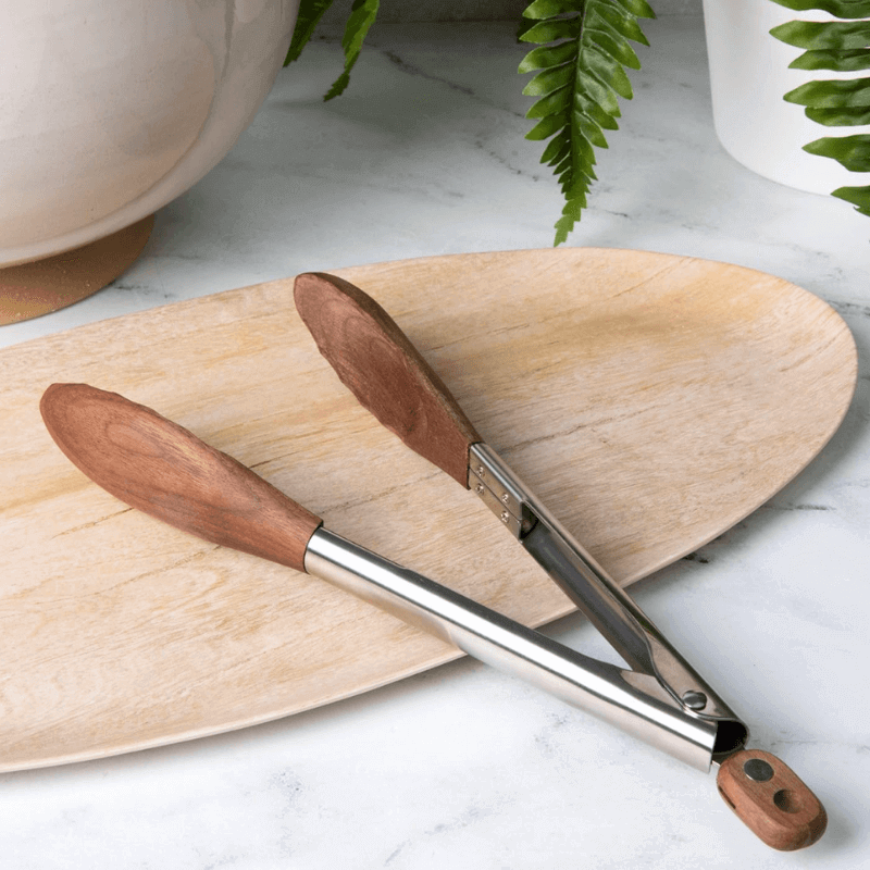 Wooden-handled tongs on a wooden board with plants in the background