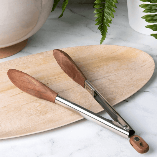Wooden-handled tongs on a wooden board with plants in the background